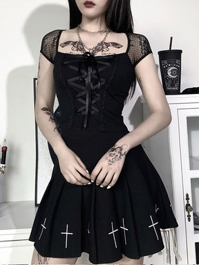 A model wears the lace-up black crop top with a pleated skirt bearing white cross motifs, standing in a room with white furniture.
