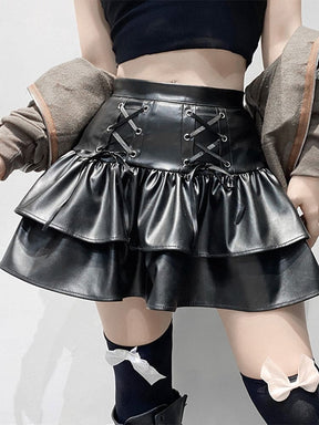 A kneeling model wears a glossy black tiered mini skirt with front ribbon lacing, a coat draped around the shoulders and bow-trimmed stockings.