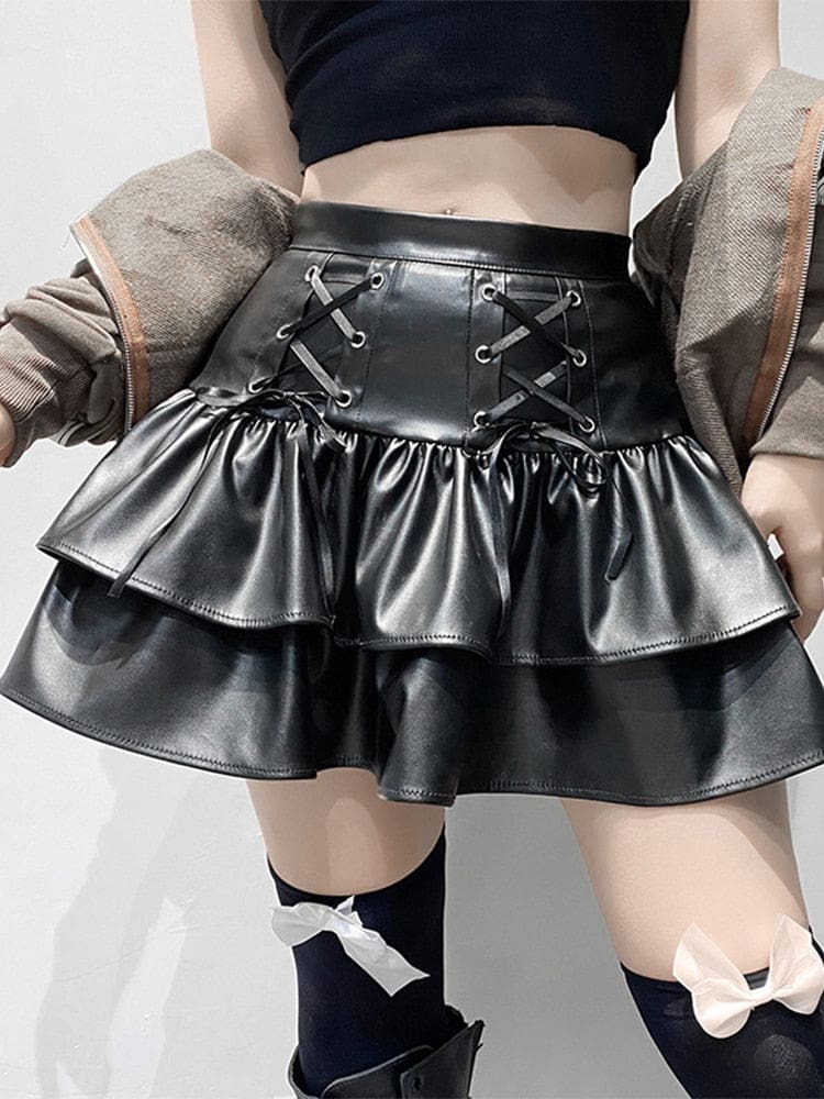 A kneeling model wears a glossy black tiered mini skirt with front ribbon lacing, a coat draped around the shoulders and bow-trimmed stockings.