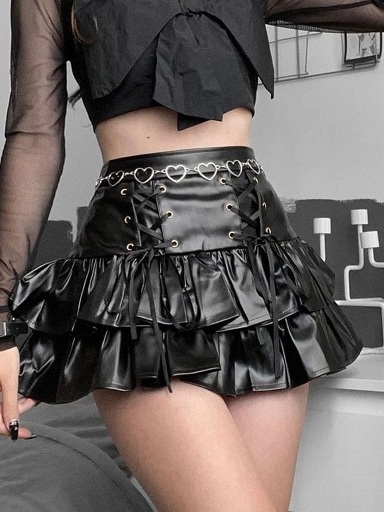 Black glossy ruffle mini skirt worn with a sheer black top and silver heart-chain belt in a bedroom. Twin front lacing panels sit above two flared tiers.