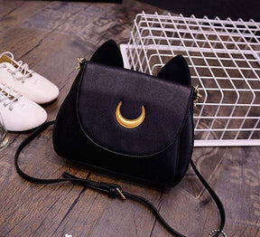 Black lunar cat purse resting on a wooden floor beside white shoes and a wire basket. Its pointed ears and gold-tone crescent stand out above the curved flap.