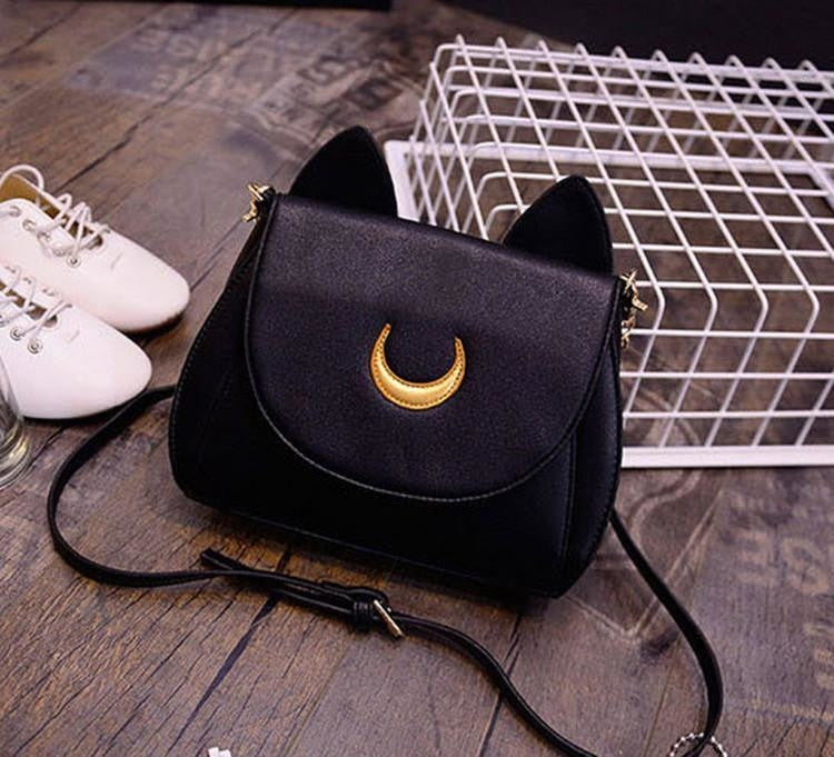 Black lunar cat purse resting on a wooden floor beside white shoes and a wire basket. Its pointed ears and gold-tone crescent stand out above the curved flap.