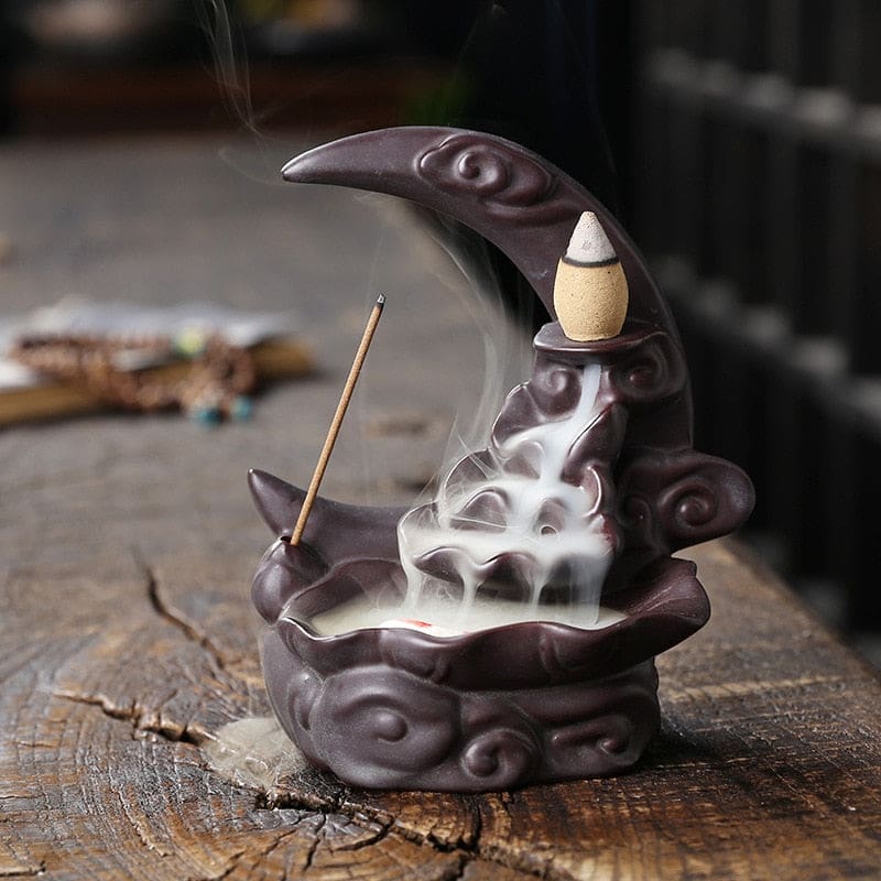 Black moon incense burner with a dark plum-toned surface, a burning cone and an upright incense stick, photographed on weathered wood.