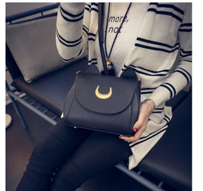 Black cat-ear crescent bag worn across a striped cardigan in a seated outfit photo, with the gold-tone moon facing forward.