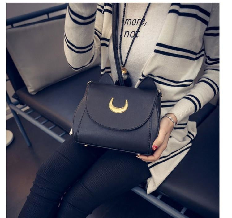 Black cat-ear crescent bag worn across a striped cardigan in a seated outfit photo, with the gold-tone moon facing forward.