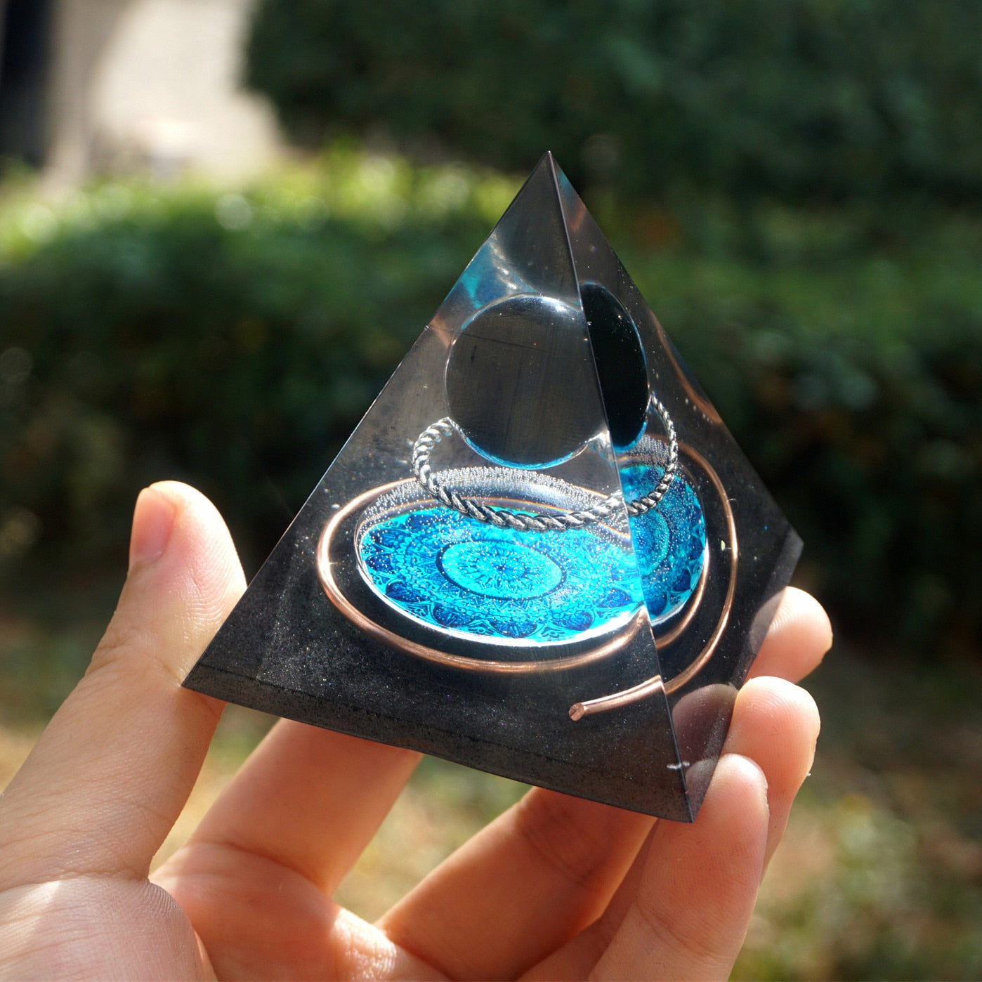 Black obsidian orgone pyramid with a dark sphere, blue mandala and copper loops. Angled view on a palm showing the circular artwork and curved wire.