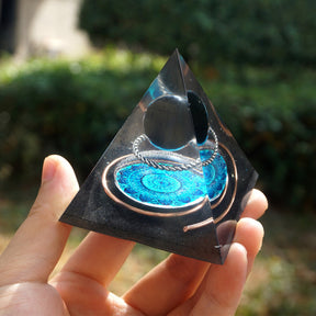 Black obsidian orgone pyramid with a dark sphere, blue mandala and copper loops. Angled view on a palm showing the circular artwork and curved wire.