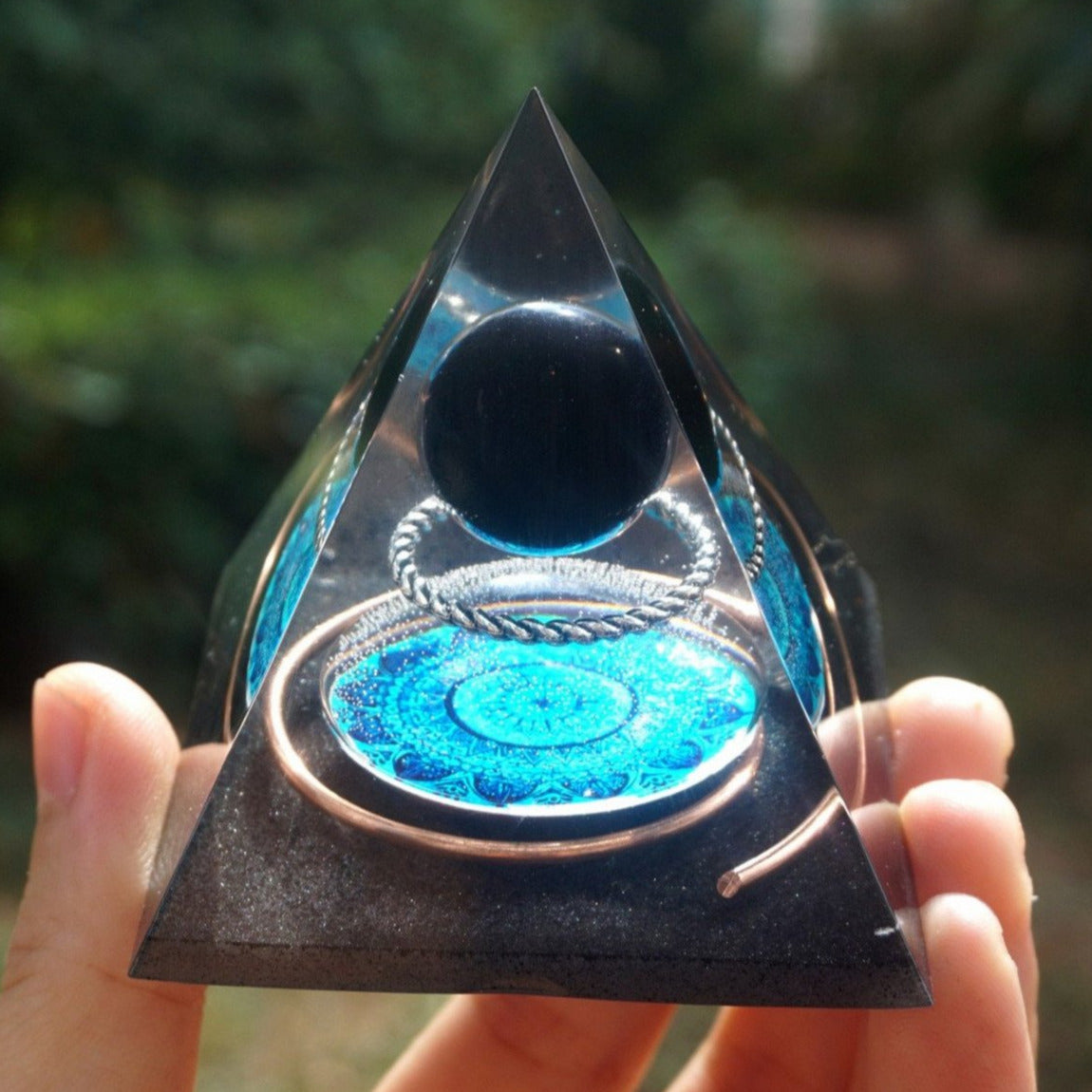 Black obsidian orgone pyramid with a dark sphere, blue mandala and copper loops. Close front view held outdoors, showing the blue disc below the sphere.