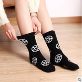 Black crew socks with repeating white pentagrams, worn on a seated model who is stretching one cuff outward. The feet rest on a light wooden floor.