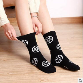 Black crew socks with repeating white pentagrams, worn on a seated model who is stretching one cuff outward. The feet rest on a light wooden floor.