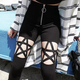 Close front view of black skinny pants with pentagram straps across open thigh panels and a silver-tone O-ring zip. Styled with a striped open shirt.