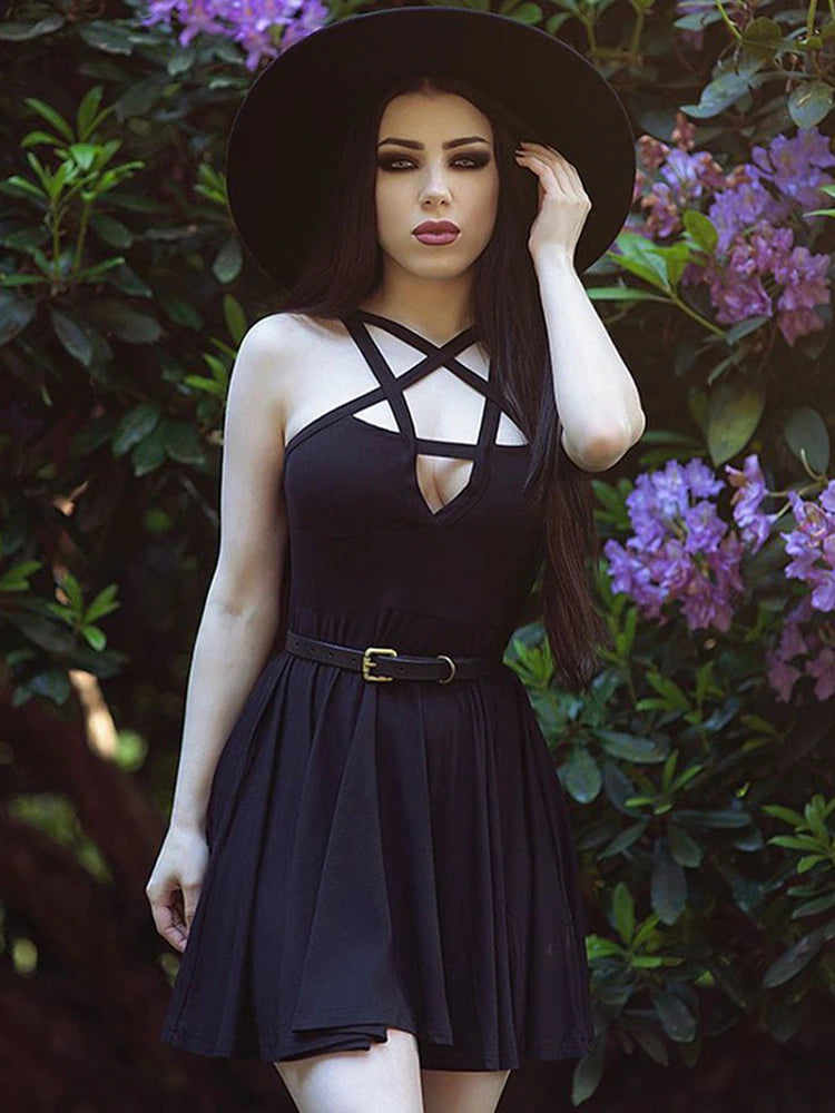 Black pentagram strap skater dress worn in a garden with a separate wide-brim hat and slim belt. Crossed neckline straps frame a front cutout above the fitted waist and flared skirt.
