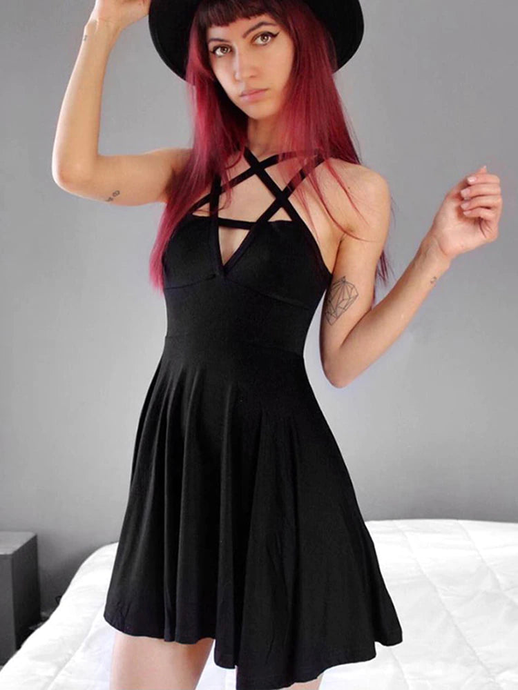 Black pentagram harness neckline dress worn beside a bed with a separate black hat, showing the crossed chest straps, front cutout and flared skirt.