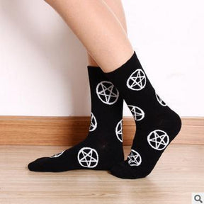 Side view of black pentagram socks on crossed ankles, showing white circled stars across the legs and feet against a pale wall and wooden floor.