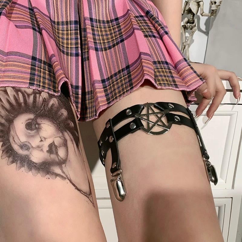 Black pentagram thigh garter seen from an angle beside a tattooed leg, with round rivets and the two hanging stocking clips visible.