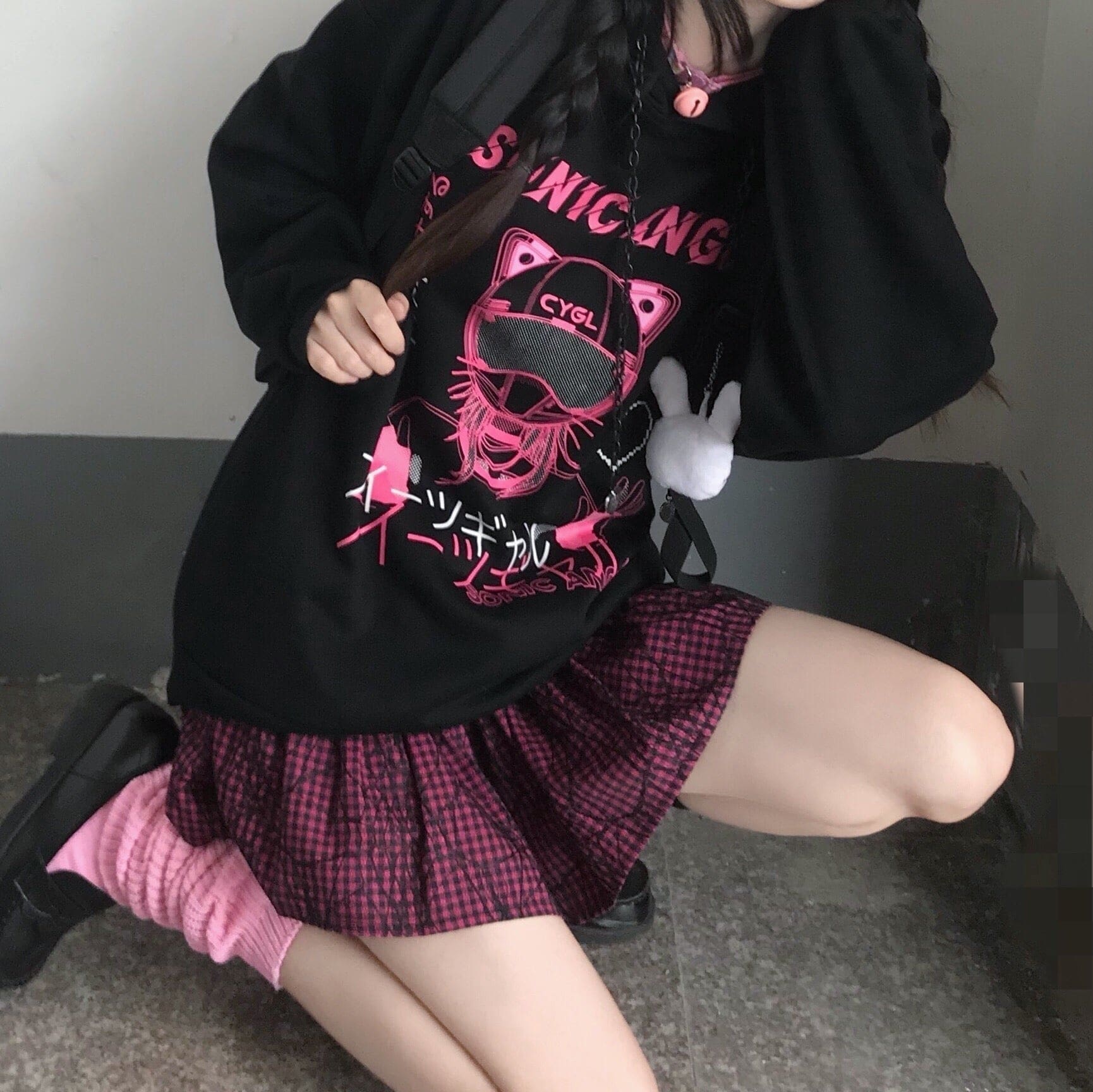 Seated view of the black graphic hoodie showing its ribbed lower hem above a pink plaid skirt.