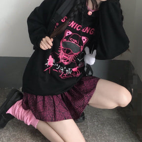 Seated view of the black graphic hoodie showing its ribbed lower hem above a pink plaid skirt.