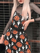 Black pumpkin-and-ghost mini dress styled over a sheer mesh top with a black belt, photographed on a blonde-haired model beside a brick wall.