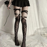 Full front view of black thigh-high rose fishnets, lace garter bands and ornate heart accents, worn beneath a black skirt against pale curtains.