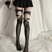 Full front view of black thigh-high rose fishnets, lace garter bands and ornate heart accents, worn beneath a black skirt against pale curtains.