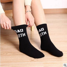 Black crew socks with bold white Sad Goth lettering near the cuffs. A seated model pulls one cuff outward above a light wooden floor.