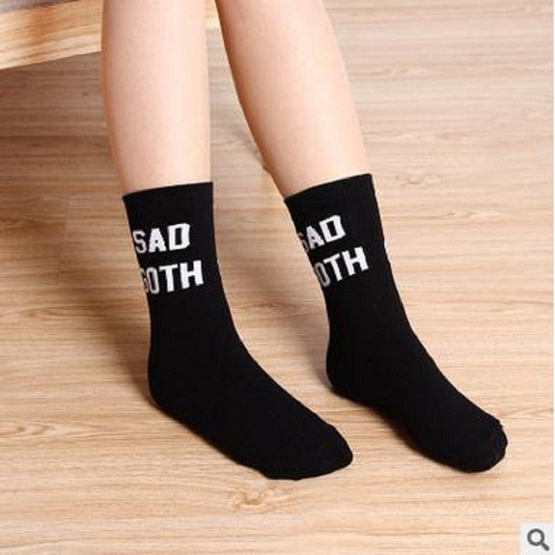 Front view of black socks worn with both feet on a wooden floor, displaying the white Sad Goth lettering across the upper cuffs.