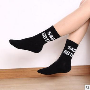 Black Sad Goth socks worn with the legs raised at an angle, showing the white cuff lettering and plain black feet against a pale wall.