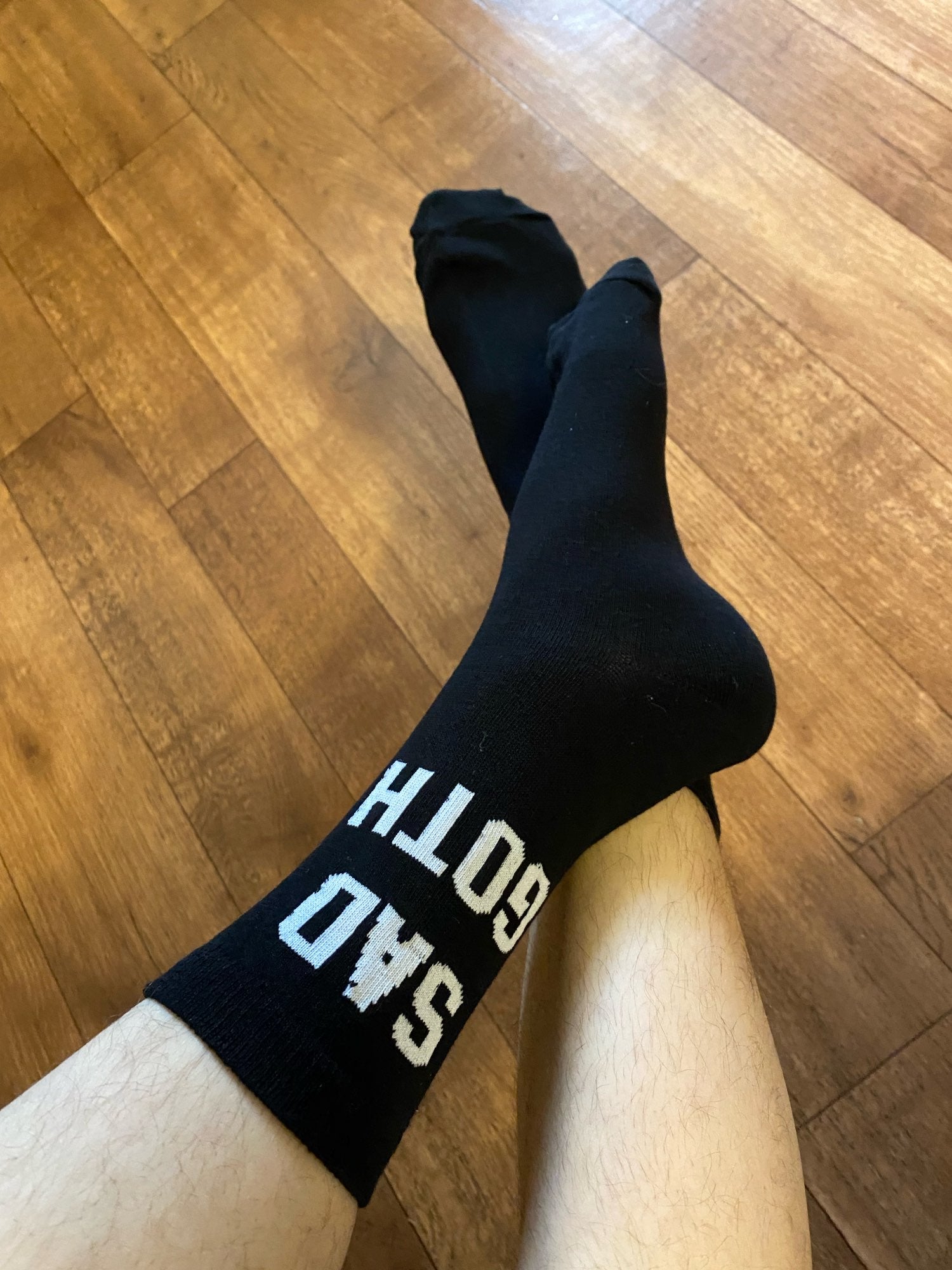 Close-up of black Sad Goth socks worn on crossed legs over a warm-toned wooden floor, with the white lettering visible near one cuff.