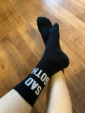 Close-up of black Sad Goth socks worn on crossed legs over a warm-toned wooden floor, with the white lettering visible near one cuff.