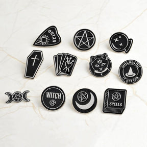Eleven separate black and silver-tone occult-inspired pin designs arranged on pale marble, including tarot cards, moons, a cat and a spellbook.