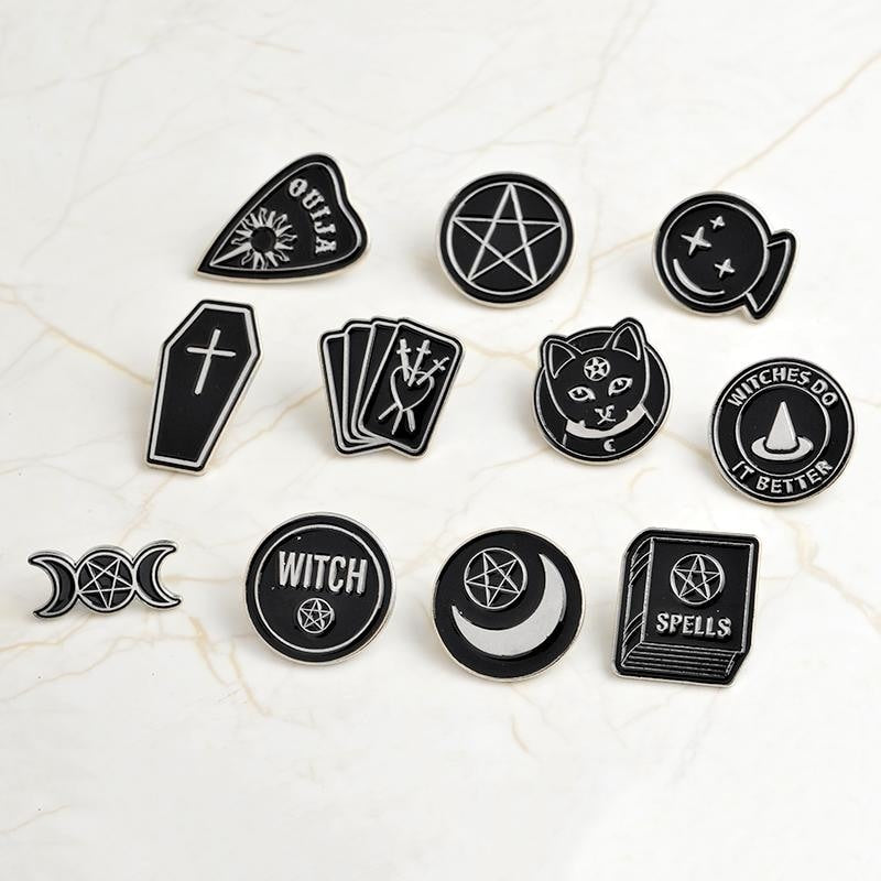 Eleven separate black and silver-tone occult-inspired pin designs arranged on pale marble, including tarot cards, moons, a cat and a spellbook.