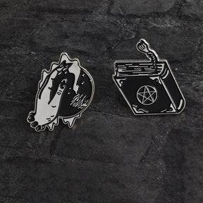 Two separate witchcraft-inspired pin designs arranged on dark textured fabric. The crystal ball and hand sit beside a pentacle spellbook with a tassel.