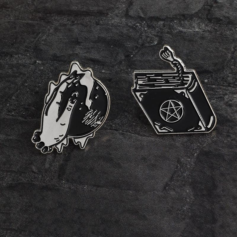 Two separate witchcraft-inspired pin designs arranged on dark textured fabric. The crystal ball and hand sit beside a pentacle spellbook with a tassel.