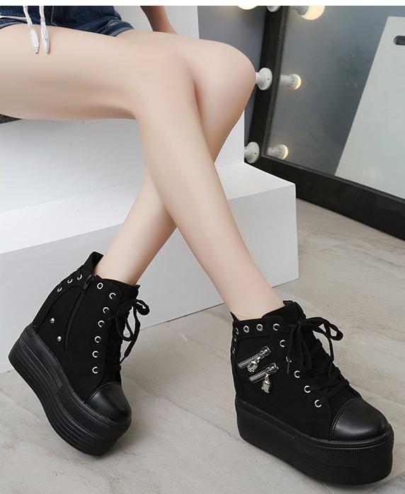 Black platform wedge sneakers worn in a seated pose, showing the front lacing, outer skull zippers and tall soles.
