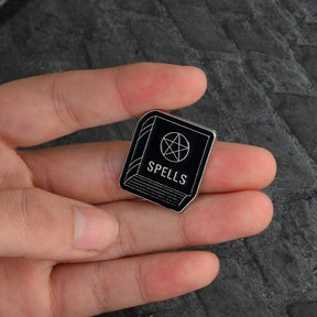 Spellbook enamel pin held between fingers, with the silver-tone word Spells and a pentagram on its black cover.