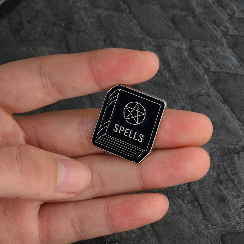 Spellbook enamel pin held between fingers, with the silver-tone word Spells and a pentagram on its black cover.