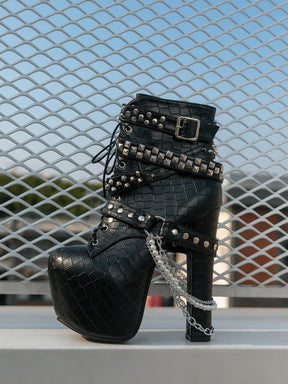 Single black reptile-textured platform boot resting against a metal grille outdoors, showing overlapping studded straps, buckles, hanging chains and a tall block heel.