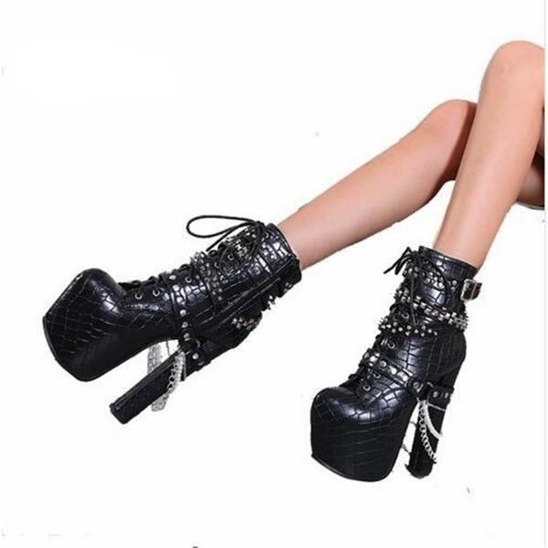 Black studded platform ankle boots worn with legs extended against white, showing front lacing, tall heels and chains hanging from the ankle straps.