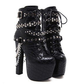 Pair of black reptile-textured platform ankle boots on white, with lace-up fronts, silver-tone studded straps, buckles and hanging chains beside tall block heels.