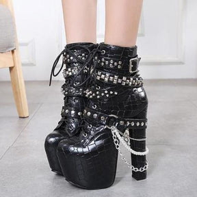 Pair of black lace-up platform ankle boots worn together on a gray tiled floor, with silver-tone studded straps and draped chains around the sides.