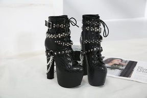 Black lace-up platform ankle boots displayed on white bedding beside an open magazine, showing their studded straps, tall heels and hanging chains.