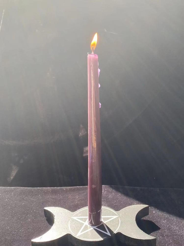 A slim pink candle burns in the black triple moon holder against a dark surface, with light falling across the crescent shapes.