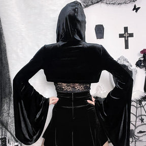 Back view of the hooded black velvet bolero with hands on hips, showing the cropped hem and wide hanging bell sleeves against a pale decorated wall.