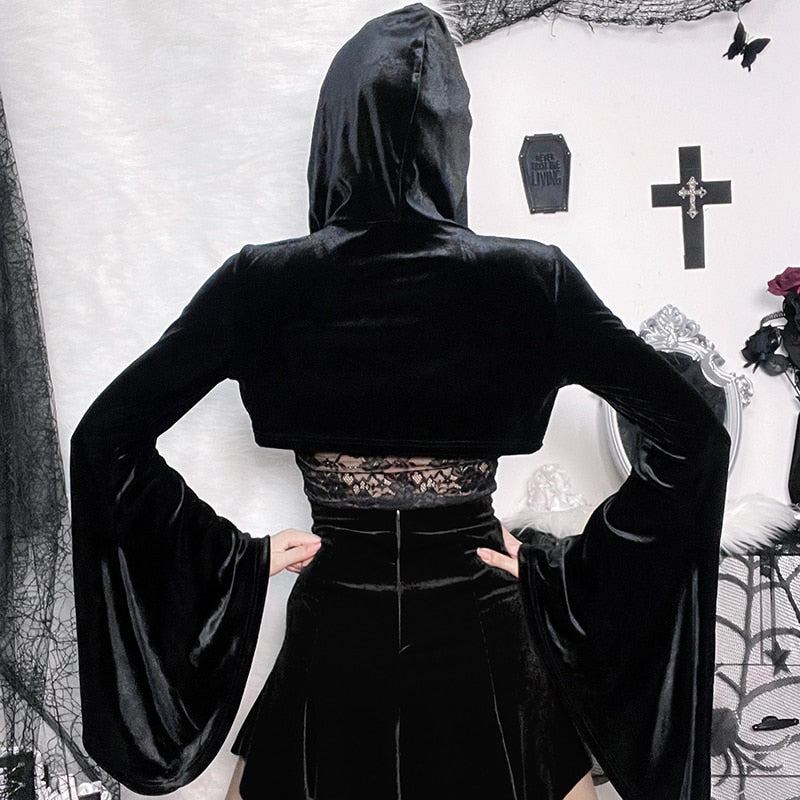 Back view of the hooded black velvet bolero with hands on hips, showing the cropped hem and wide hanging bell sleeves against a pale decorated wall.