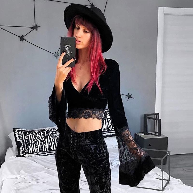 A red-haired model in a wide-brim black hat photographs the velvet and lace crop top in a bedroom mirror, wearing dark patterned trousers.