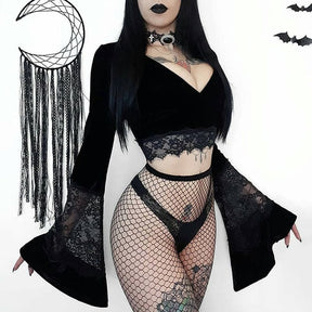 Front outfit view of the black velvet crop top with both flared sleeves spread outward, styled with fishnets against a pale wall.