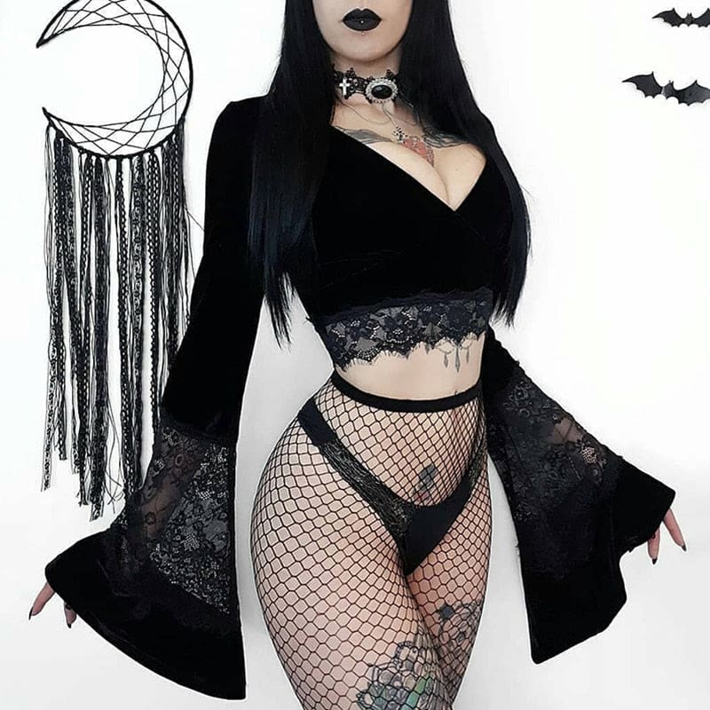 Front outfit view of the black velvet crop top with both flared sleeves spread outward, styled with fishnets against a pale wall.