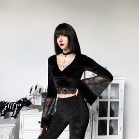 Three-quarter front pose in the black velvet bell-sleeve crop top, styled with pinstripe trousers beside white cabinets.