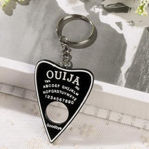 Black planchette resin keychain with a white border, alphabet and numbers, displayed on lace with its silver-tone ring resting on an open book.