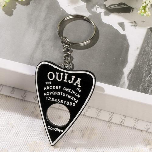 Black planchette resin keychain with a white border, alphabet and numbers, displayed on lace with its silver-tone ring resting on an open book.